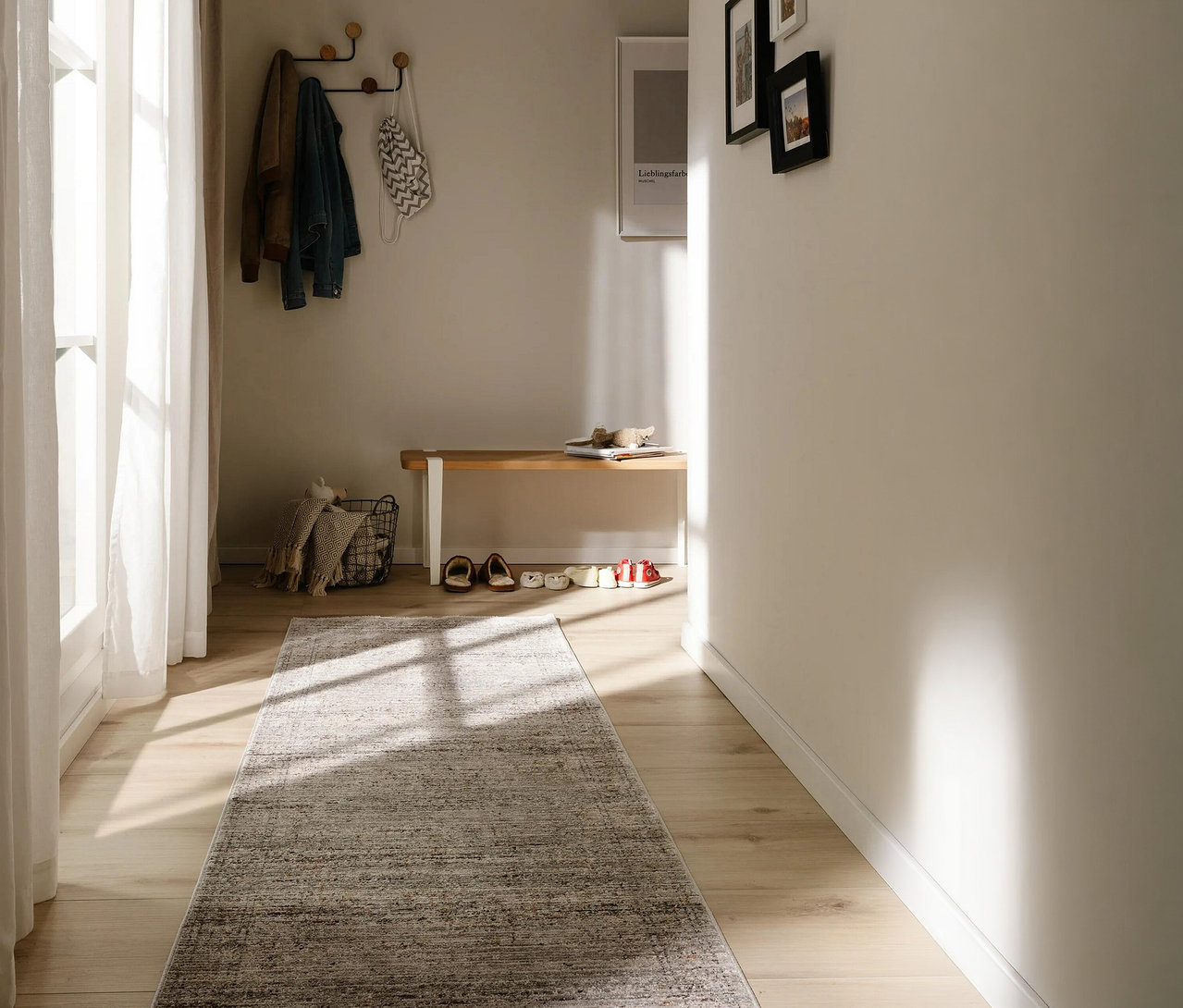 Heller Flur mit benuta Läufer »Anwar« in Grau-Blau auf Holzboden. Sonnenlicht fällt durch ein Fenster. Eine Garderobe mit Jacken hängt über einer Holzbank mit Schuhen und Korb.