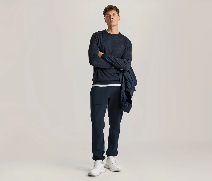 Mann in dunkelblauem Langarmshirt, dunkelblauer Hose und weißen Sneakern, mit einer dunkelblauen Jacke über dem Arm, steht mit verschränkten Armen.