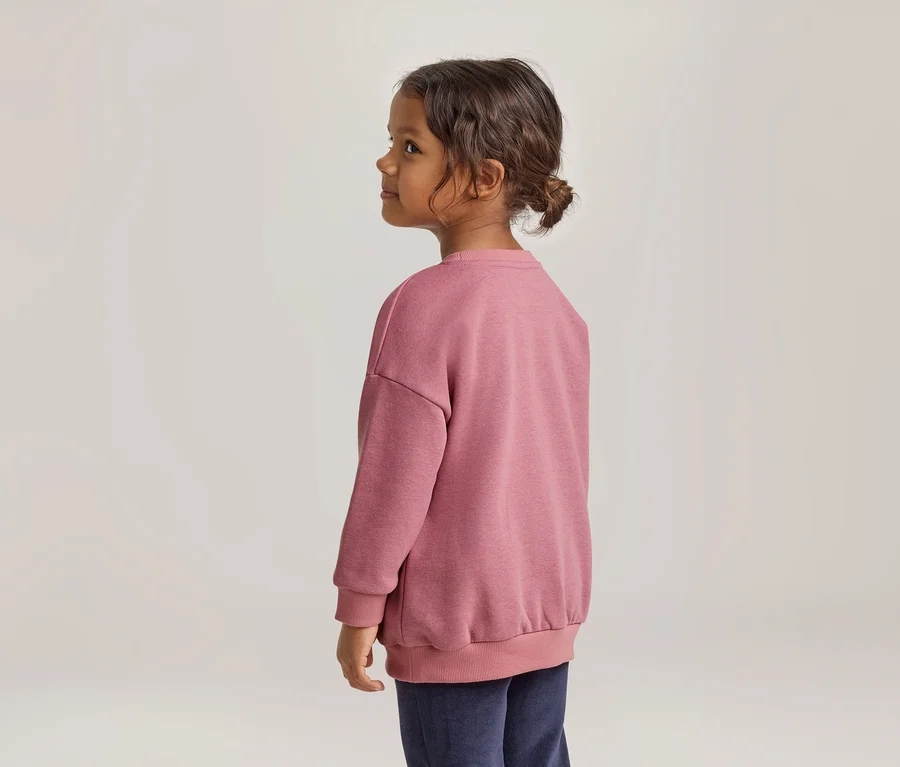 Junges Kind mit dunklem, lockigem Haar, das zu einem Dutt gebunden ist, von der Seite gesehen, trägt ein pinkes Sweatshirt und dunkle Hose, blickt nach links.