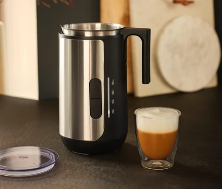 Ein Milchaufschäumer aus Edelstahl steht neben einem Glas Kaffee mit Milchschaum.