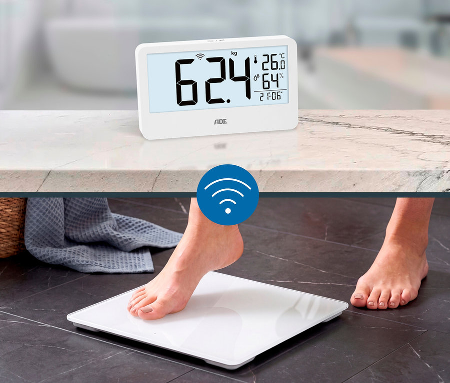 Ein weißes Digitaldisplay zeigt 62,4 kg, Temperatur, Luftfeuchtigkeit und Uhrzeit auf einer Arbeitsplatte. Darunter treten nackte Füße auf eine weiße Digitale Personenwaage auf dunklem Boden, verbunden durch ein WLAN-Symbol.