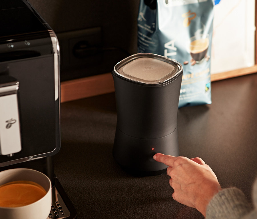 Eine Frau schaltet einen anthrazitfarbenen elektrischen Milchaufschäumer ein. Daneben stehen eine Kaffeemaschine und eine Tasse Kaffee.