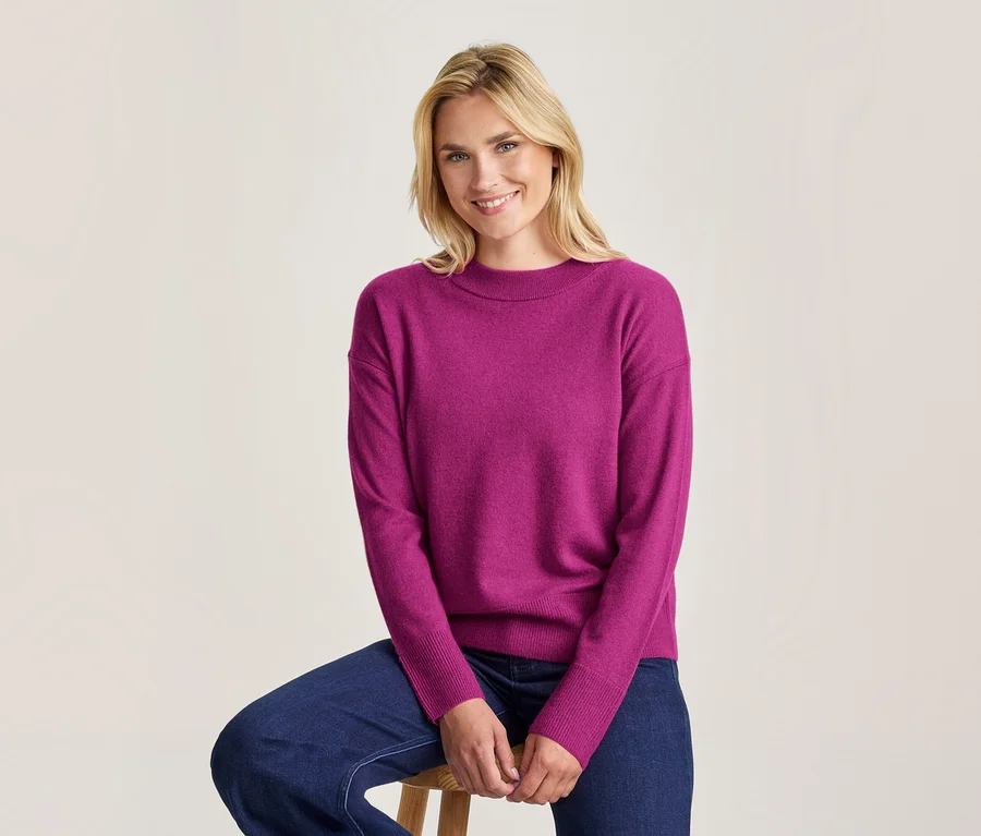 Lächelnde blonde Frau sitzt auf einem Holzhocker und trägt einen magentafarbenen Langarmpullover und dunkelblaue Jeans.