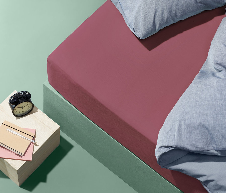 Ein rosafarbenes Biber-Spannbettlaken in Normalgröße liegt auf einem Bett mit blauen Kissen und einer Bettdecke. Auf dem Nachttisch liegen ein Wecker und ein Notizbuch.