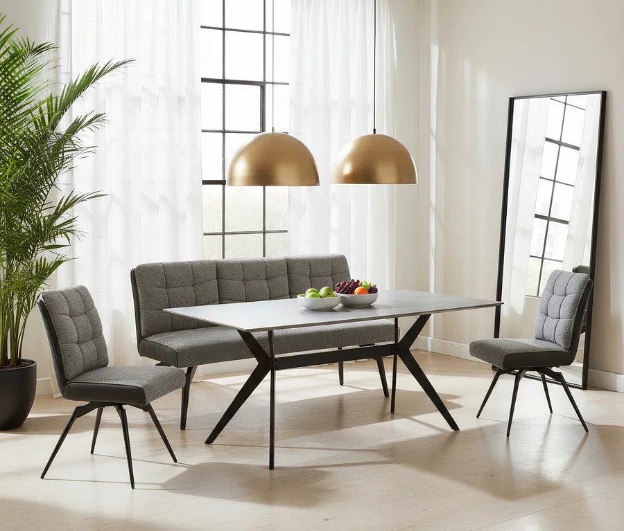 Modernes Esszimmer mit einem rechteckigen Tisch mit grauer Platte und schwarzen Kreuzbeinen. Eine graue, gesteppte Sitzbank und zwei passende Stühle umgeben den Tisch. Zwei goldene Hängelampen leuchten von oben, während auf dem Tisch Obstschalen stehen. Ein großer Spiegel und eine Pflanze schmücken den Raum.