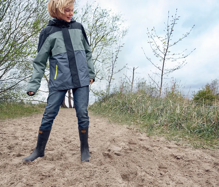 Ein Junge steht auf einem Sandweg in einer Allwetterjacke aus recyceltem Material und Jogg-Denim und trägt Gummistiefel.