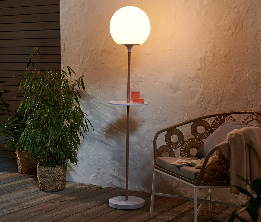 Eine LED-Stehleuchte mit Tisch steht neben einem Rattansessel auf einer Holzterrasse.