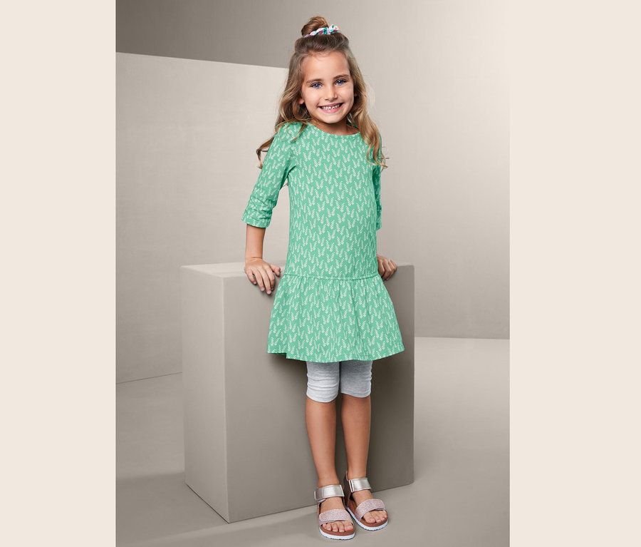 Mädchen in grünem Jerseykleid mit Blumenmuster, steht neben einem grauen Würfel, trägt graue Leggings und Sandalen.