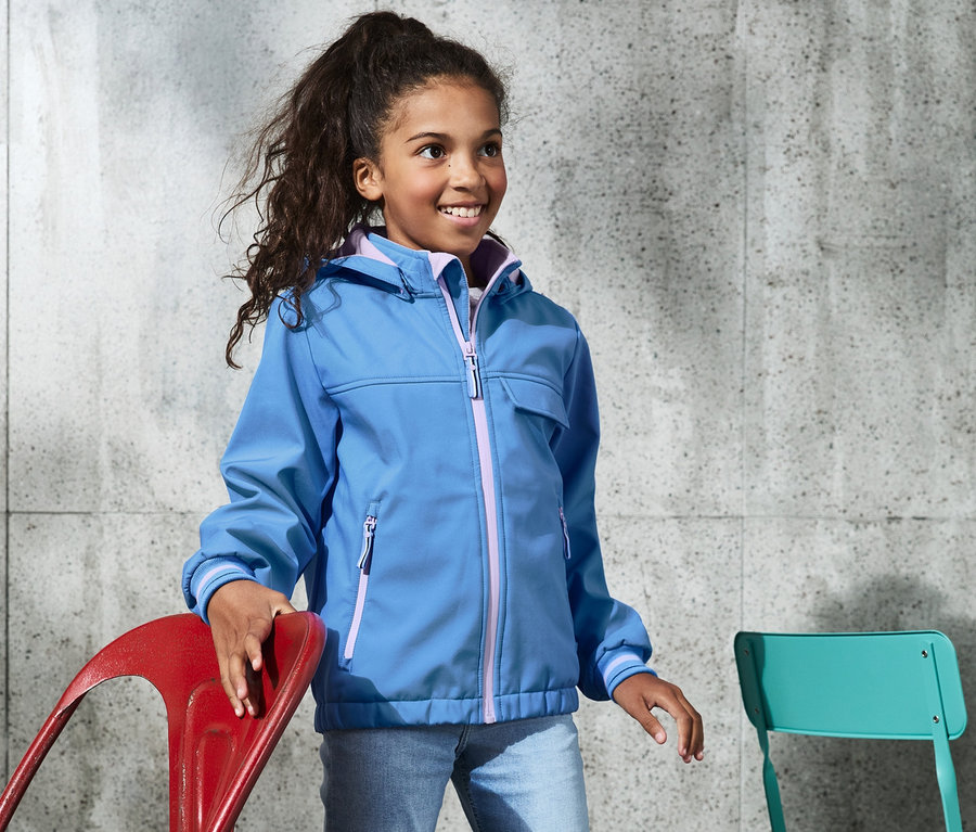 Mädchen trägt eine blaue Softshelljacke mit recyceltem Material und steht.