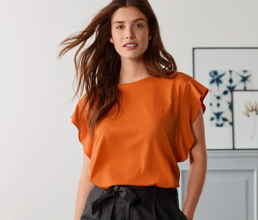 Frau posiert in orangefarbenem Shirt mit Volant und schwarzen Shorts aus Leinenmix.