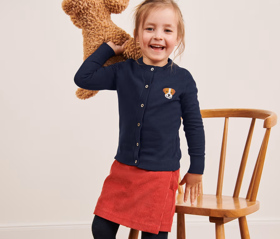Ein lächelndes Mädchen posiert mit einem Teddybären auf der Schulter. Sie trägt eine Kleinkinder-Strickjacke und einen roten Rock.