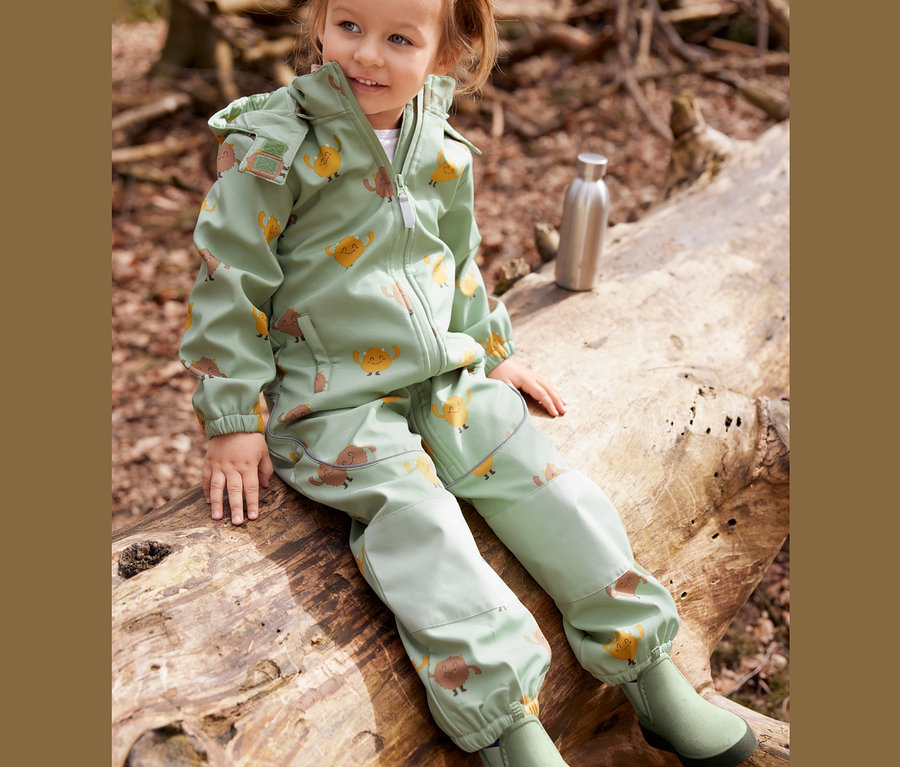 Mädchen sitzt auf Baumstamm und trägt einen grünen Softshell-Einteiler mit Monsterprint.