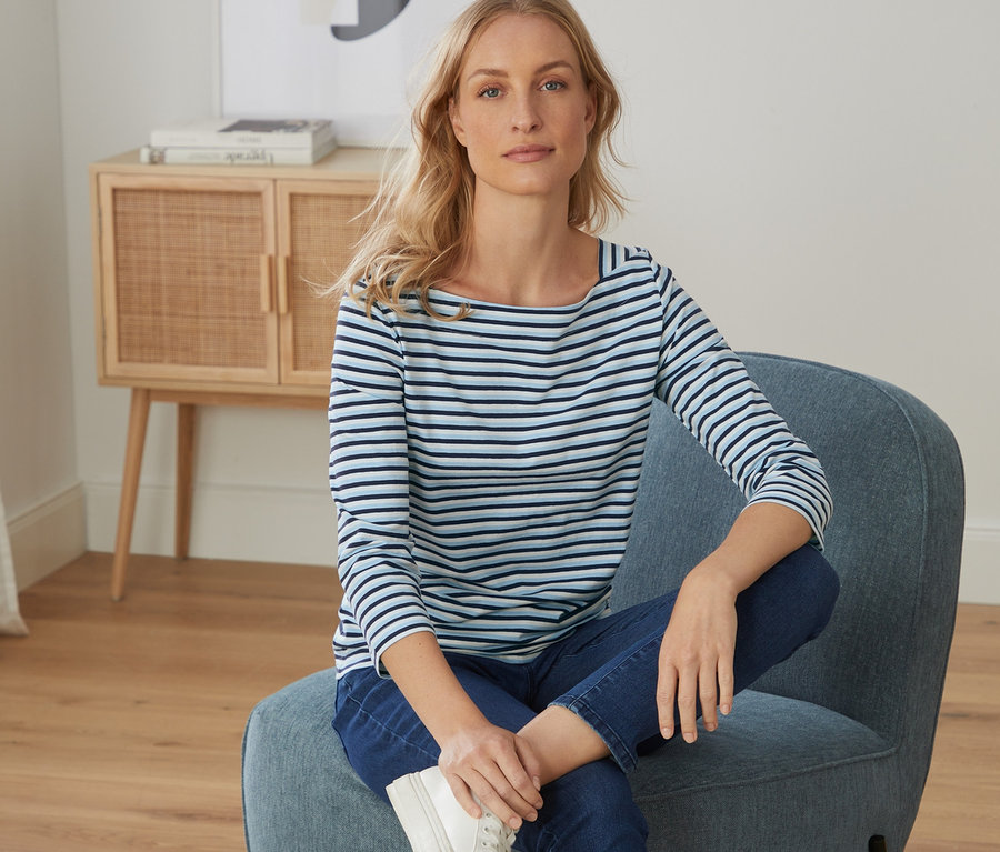 Blonde Frau sitzt im Sessel und trägt ein hellblau-dunkelblau-weiß gestreiftes Shirt mit 3/4-Arm, Jeans und Turnschuhe.