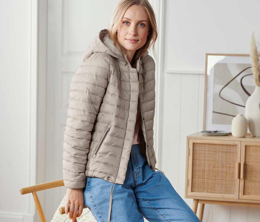 Frau sitzt auf einem Stuhl und trägt eine Steppjacke mit Kapuze.