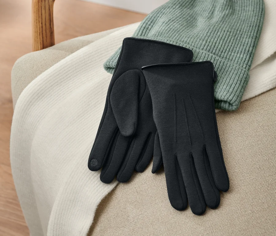 Schwarze Sweathandschuhe liegen auf einem beigen Stuhl neben einer grünen Mütze.