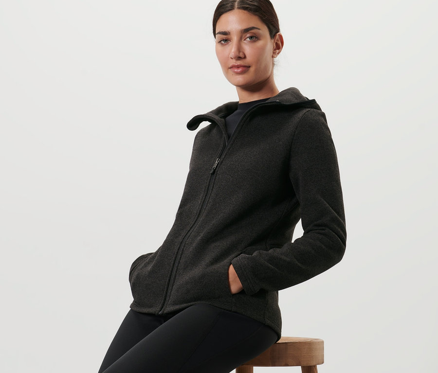 Frau sitzt auf einem Hocker und trägt eine schwarz melierte Strickfleece-Jacke.