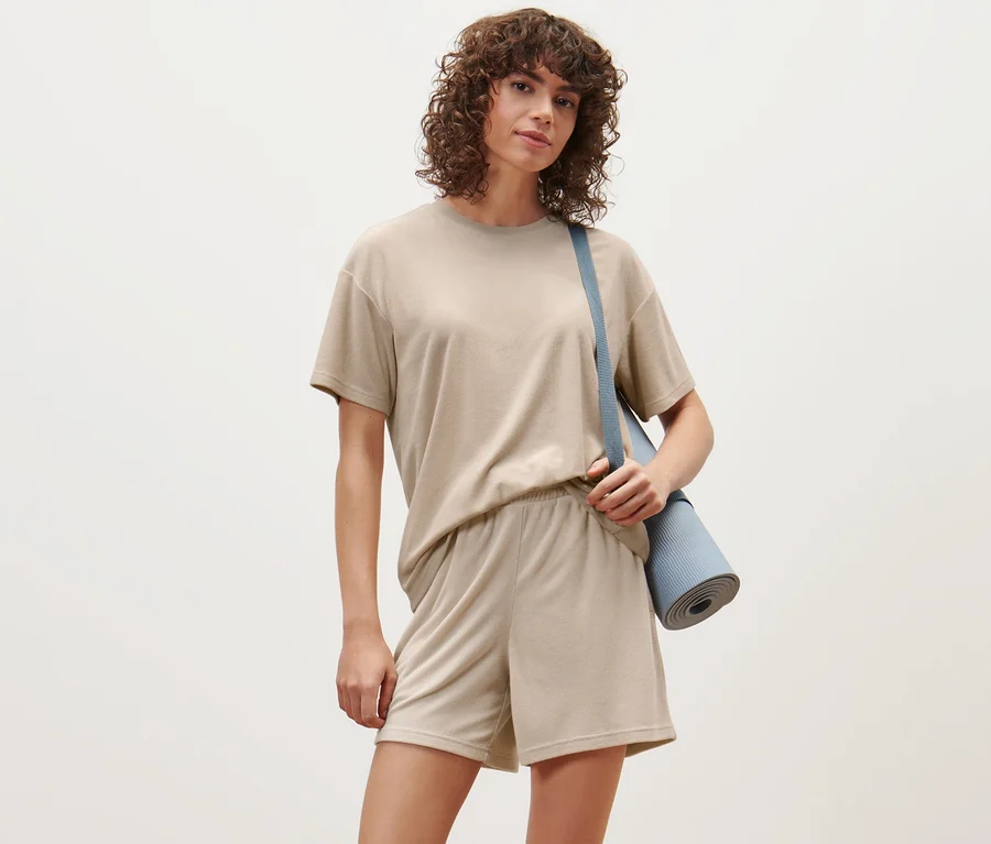 Frau trägt ein Frottier-Shirt und Shorts, eine blaue Yogamatte hängt über ihrer Schulter.