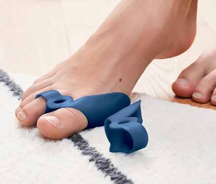 Fuß mit Hallux-Valgus-Schutz auf einem Teppich, im Hintergrund ein Paar Schuhe.