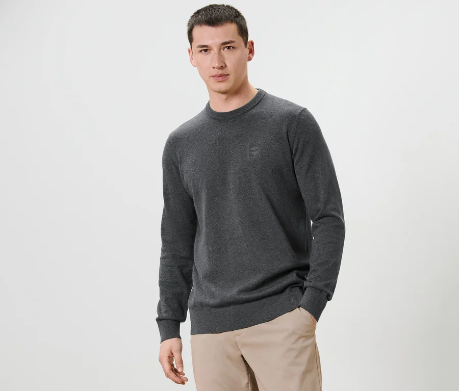 Mann posiert vor einem weißen Hintergrund und trägt einen grauen Pullover mit Rundhalsausschnitt und eine beige Hose.