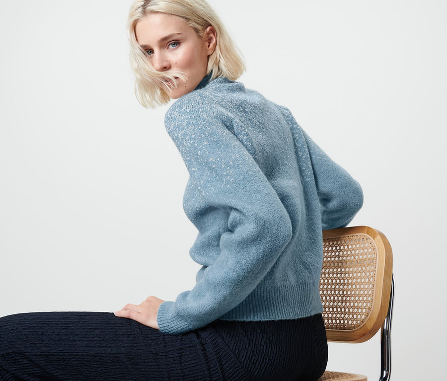 Blonde Frau sitzt auf einem Stuhl und trägt einen blauen Pullover mit Glitzerstrick.