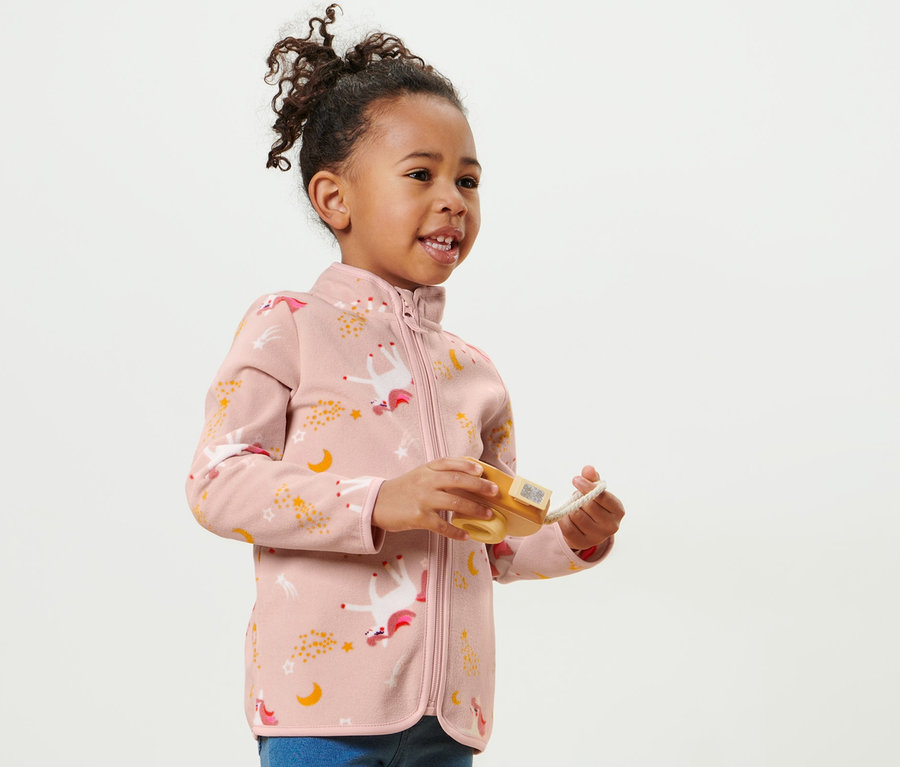 Lächelndes Mädchen trägt eine Kinder-Fleecejacke mit Einhorn-Motiv und spielt mit einem Spielzeug.