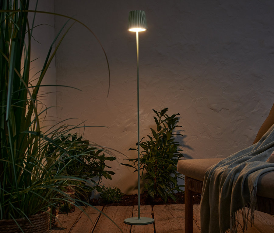 Eine salbeifarbene LED-Akku-Stehleuchte beleuchtet eine Terrasse mit Pflanzen und einer Liege.