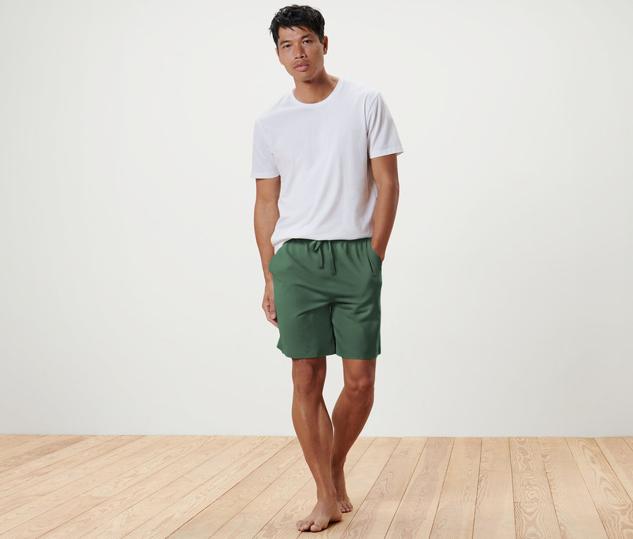 Mann posiert auf Holzboden in olivgrünen Relaxshorts und weißem T-Shirt.