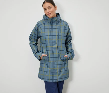 Eine Frau steht mit den Händen in den Taschen einer blau karierten Regenjacke.