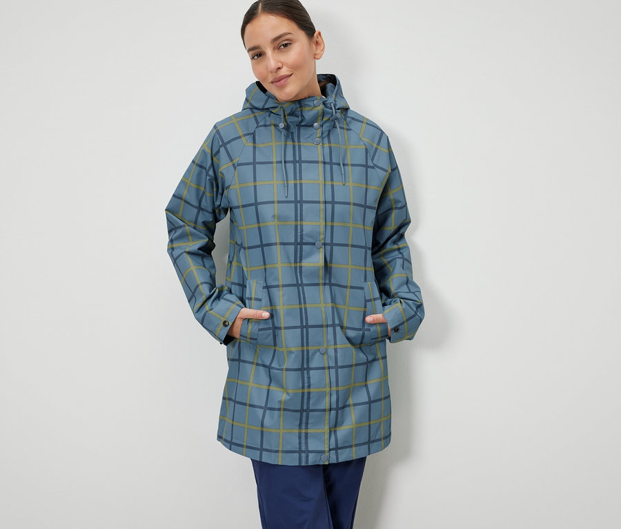 Eine Frau steht mit den Händen in den Taschen einer blau karierten Regenjacke.