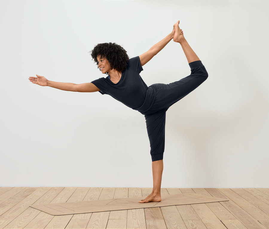 Frau in schwarzer Sportbekleidung steht in Tänzerpose auf einer Yogamatte.