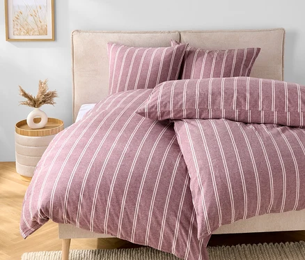 Schlafzimmer mit einem Bett mit lila-weiß gestreifter Bettwäsche, zwei Kissen und einem beigen Kopfteil. Neben dem Bett steht ein Tisch mit einer Vase.