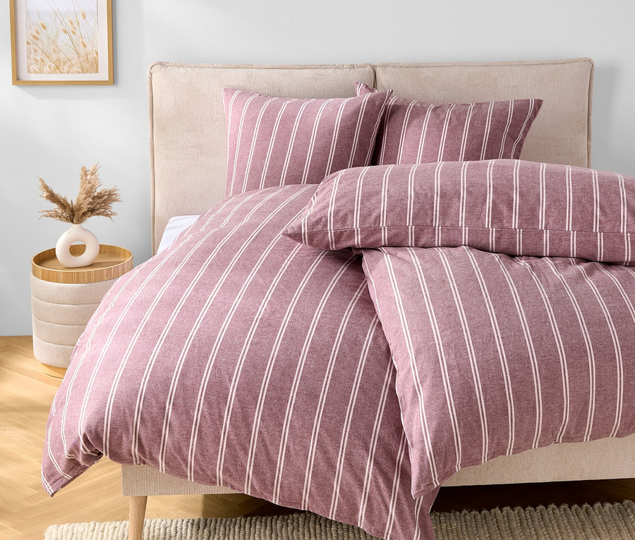 Schlafzimmer mit einem Bett mit lila-weiß gestreifter Bettwäsche, zwei Kissen und einem beigen Kopfteil. Neben dem Bett steht ein Tisch mit einer Vase.