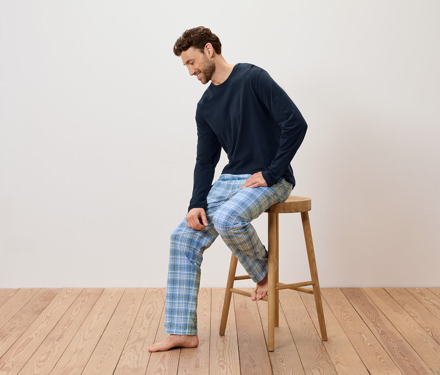 Mann sitzt auf einem Holzhocker, trägt einen dunkelblauen Schlafanzug und eine blau karierte Hose.