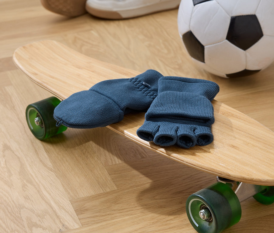 Blaue Fingerhandschuhe liegen auf einem hölzernen Skateboard mit grünen Rädern. Im Hintergrund befinden sich ein Fußball und Schuhe.