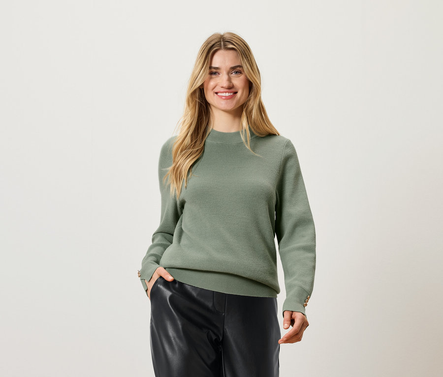 Frau mit blonden Haaren posiert in einem olivgrünen Pullover und schwarzen Lederhosen.