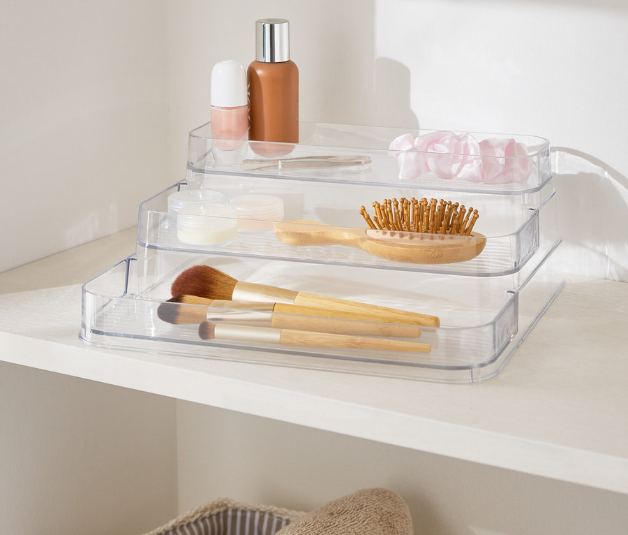 Dreistufiger, durchsichtiger Make-up-Organizer auf einem weißen Regal neben Körben mit Handtüchern.