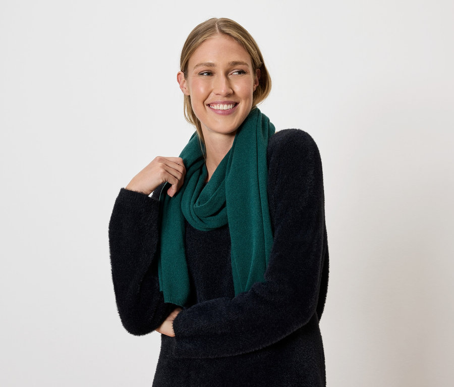 Frau mit blonden Haaren und einem Lächeln posiert vor einem hellen Hintergrund mit einem dunkelgrünen Schal und Pullover.