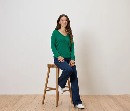 Eine Frau sitzt auf einem Holzhocker und trägt einen grünen Pullover und Jeans.