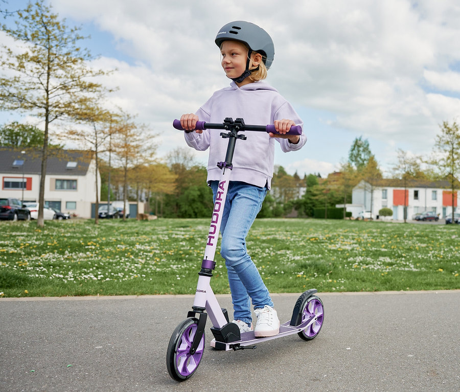 Ein Mädchen fährt mit einem lavendelfarbenen HUDORA BigWheel® 205 advanced Roller und einem grauen Helm auf einer asphaltierten Straße.