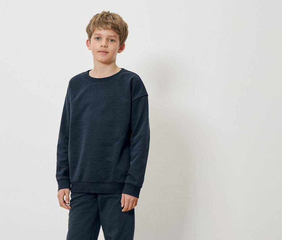 Ein Junge steht und trägt einen dunkelblauen Pullover und eine Hose.