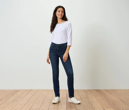 Eine Frau in einer weißen Bluse und dunkelblauen Jeans posiert auf einem Holzboden.