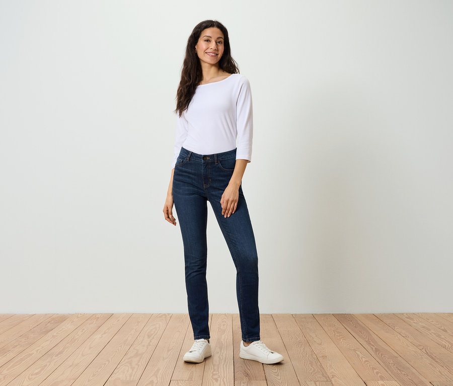 Eine Frau in einer weißen Bluse und dunkelblauen Jeans posiert auf einem Holzboden.