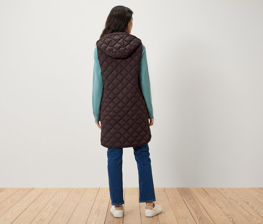 Eine Frau steht mit dem Rücken zur Kamera und trägt eine braune Steppweste mit Kapuze, einen blauen Pullover und Jeans.