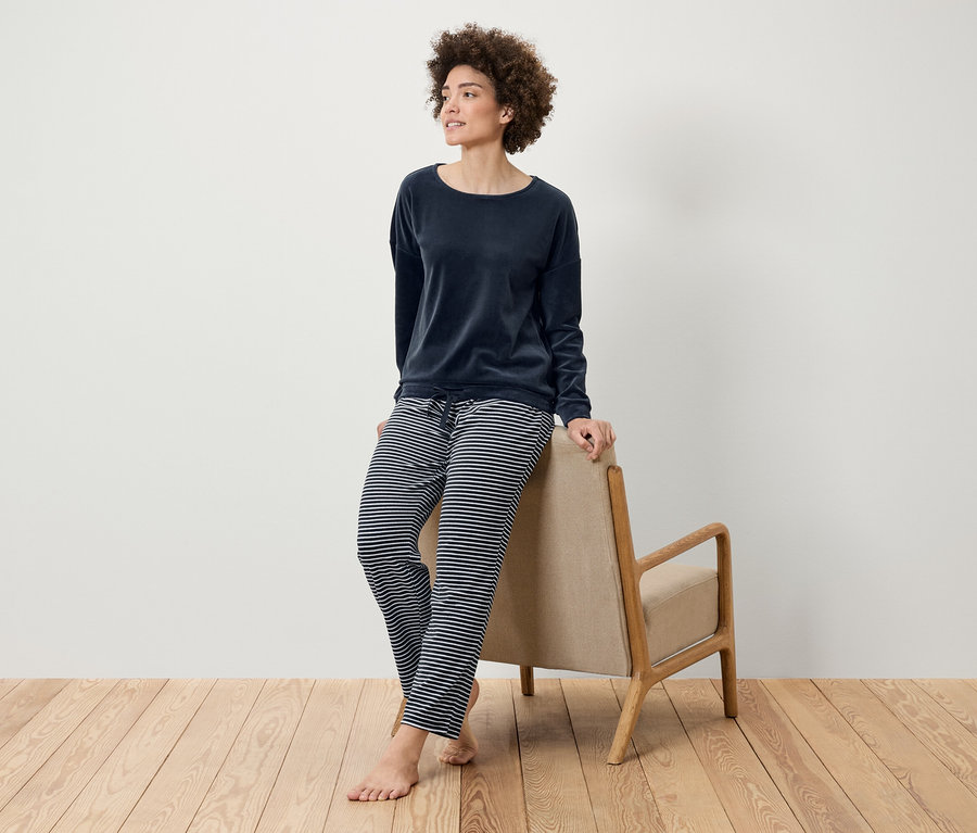 Frau mit Afro-Frisur posiert in einem marineblauen Samttop und einer gestreiften Hose, die sich an einen beigen Sessel auf einem Holzboden lehnt.