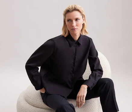 Blonde Frau sitzt auf einem beigen Sessel und trägt ein schwarzes Hemd und eine Hose.