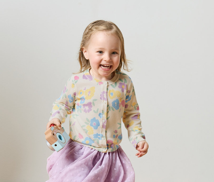 Lächelndes Mädchen mit blonden Haaren, gebundenen Zöpfen, geblümter Strickjacke und lila Rock hält eine Spielzeugkamera.