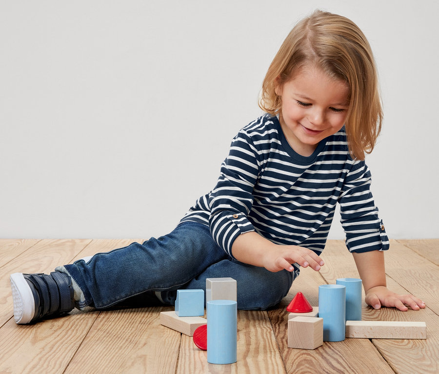 Kind sitzt am Boden und spielt mit bunten Holzklötzen. Es trägt ein 2 Kinder-Langarmshirts.