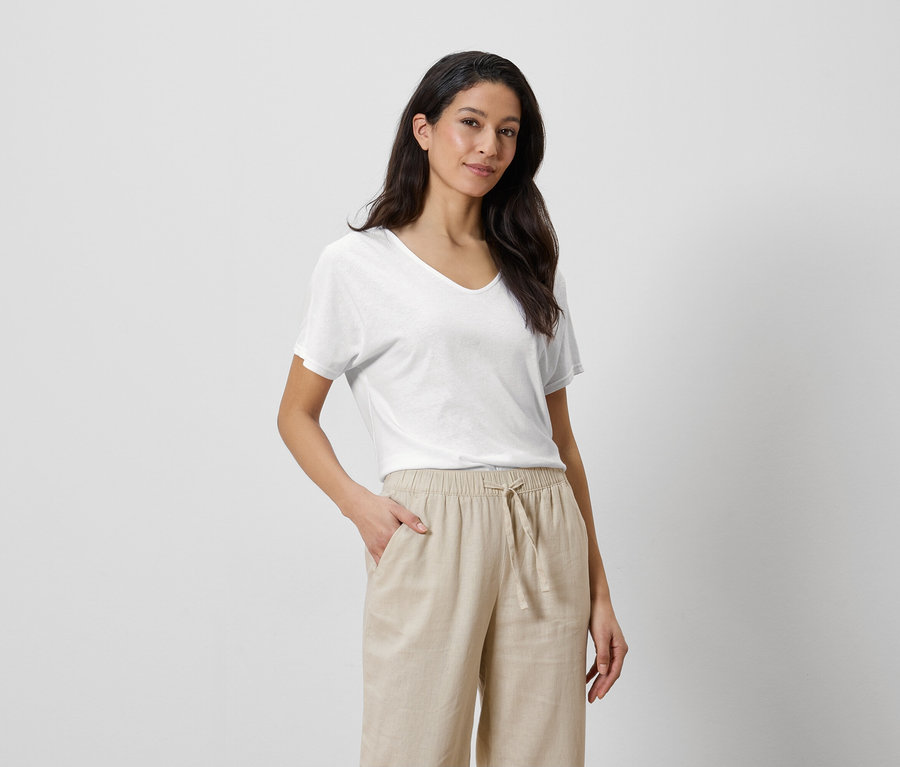 Frau mit weißem T-Shirt und beigen Hosen, eine Hand in der Hosentasche.