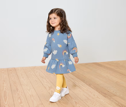 Ein kleines Mädchen mit lockigem Haar trägt ein blaues Kleid mit Regenbogenmuster, gelbe Leggings und weiße Schuhe.