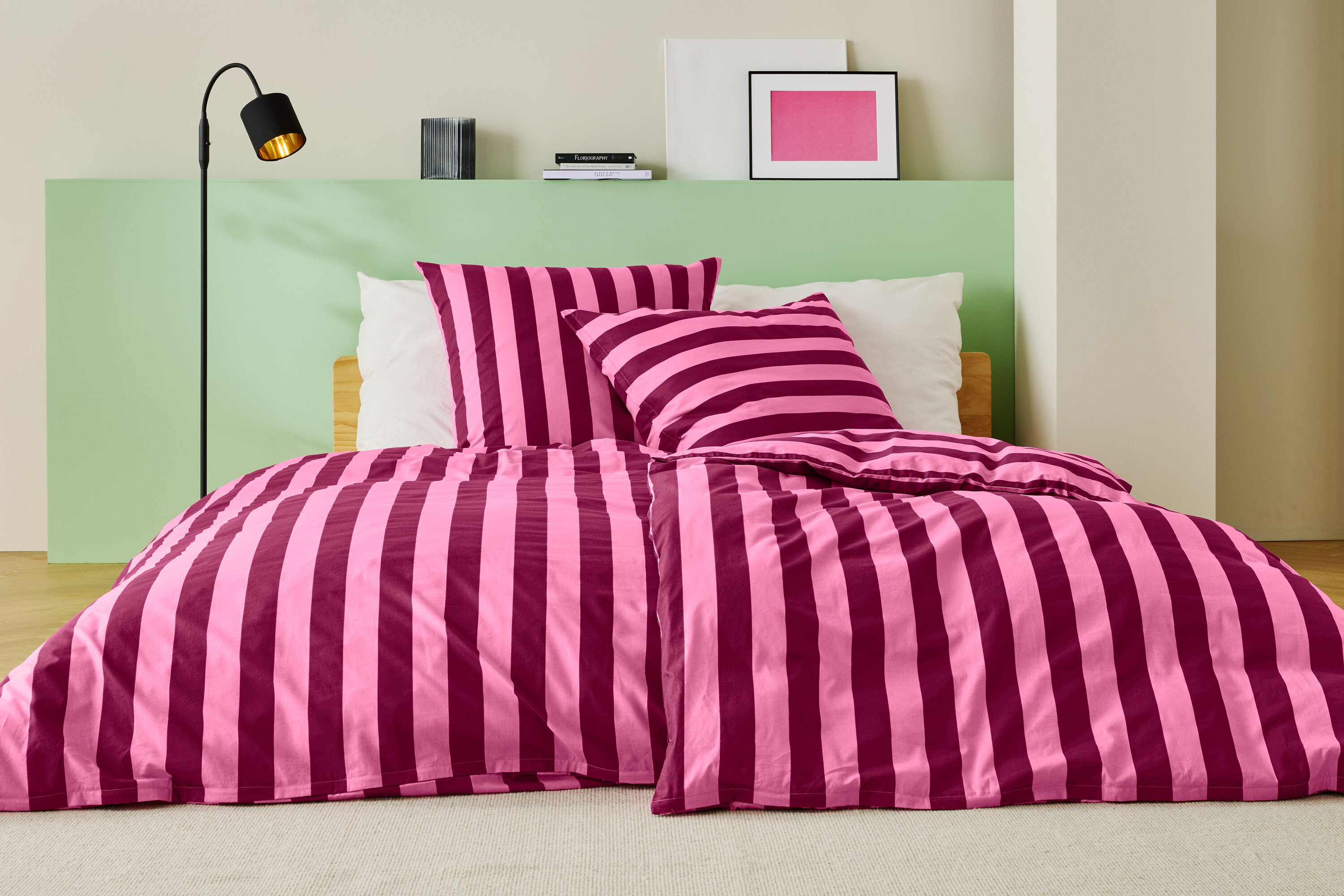 Bett mit rosa-weinroter Streifenbettwäsche, zwei Kissen und einer Stehlampe.